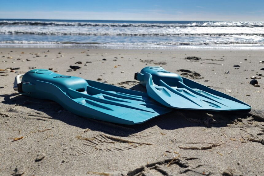 Fourth Element Tech fins lying flat on the beach sand with waves breaking behind