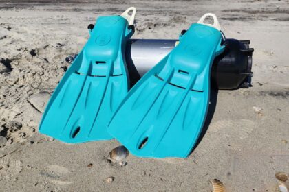 Teal Fourth Element Tech fins leaning against a scuba cylinder on the sand at the beach test site