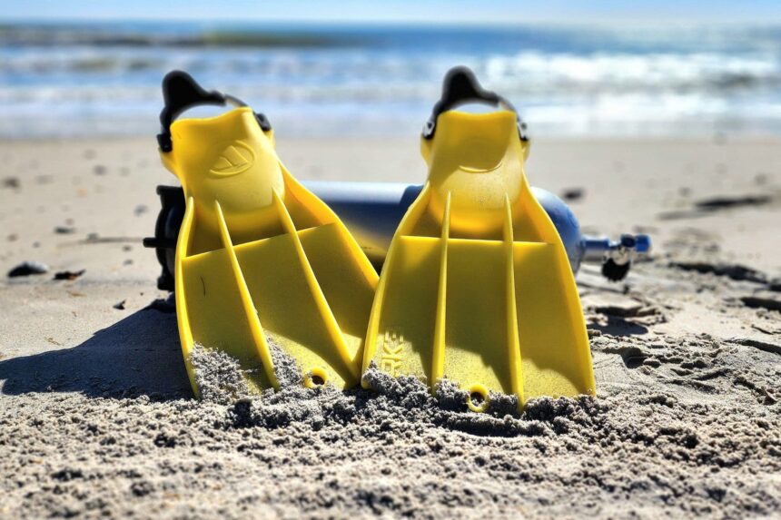 Yellow Apeks RK3 fins standing upright in beach sand with a scuba cylinder behind them