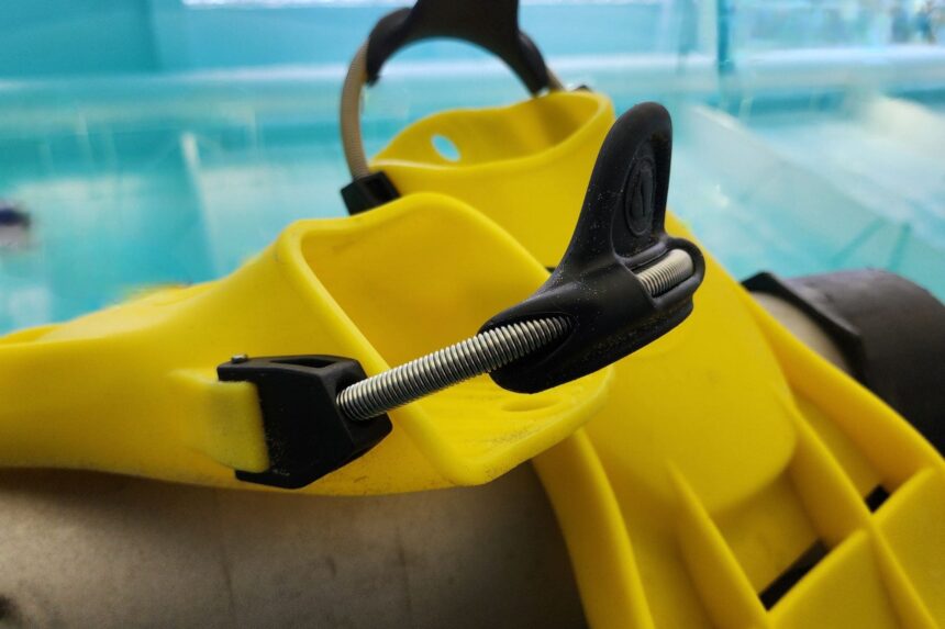 Close-up of the stainless spring strap and heel notch on a yellow Apeks RK3 fin beside a pool
