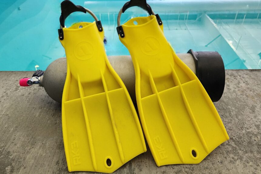 Two yellow Apeks RK3 fins propped against a scuba cylinder at the edge of a pool