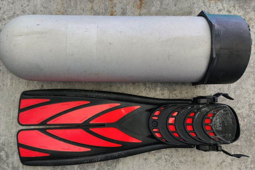 Black and red Atomic Aquatics SplitFin laid out beside an aluminum scuba cylinder on concrete