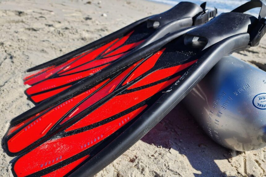 Black and red Atomic Aquatics split fins lying on beach sand next to a scuba cylinder