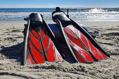 Atomic Aquatics split fins in red and black standing in the sand beside a scuba cylinder