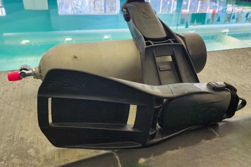 Two ScubaPro S-Tek fins leaning on a scuba cylinder on the pool deck, vented blades facing out