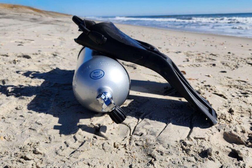 ScubaPro S-Tek fin propped against an aluminum cylinder in the sand with surf behind