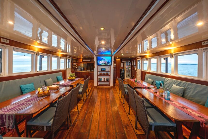 Dining saloon aboard the Ambai with long tables, a TV, and window views of the sea