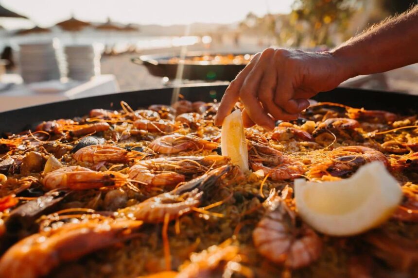 Chef adding a lemon wedge to a large seafood paella on the beach