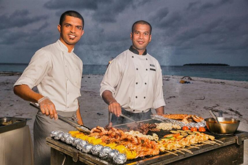 Two chefs grilling seafood and skewers on the beach at sunset