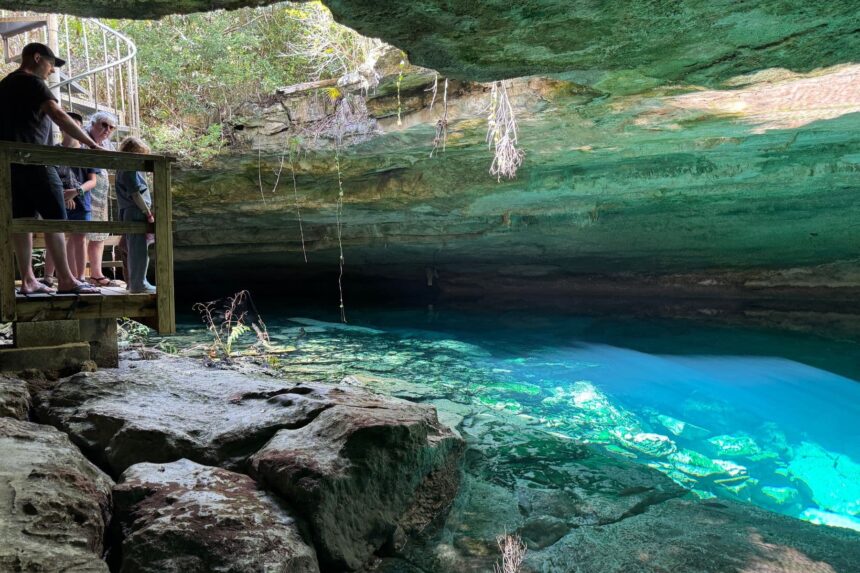 Visitors viewing a clear blue cenote pool from a wooden platform in Lucayan National Park