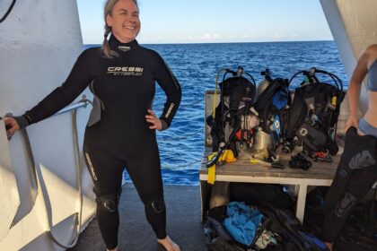 DIVEIN tester wearing the Cressi Otterflex wetsuit on a dive boat deck with rigged BCDs behind her