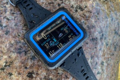 Shearwater Peregrine dive computer with blue bezel resting wet on a rock showing its surface screen