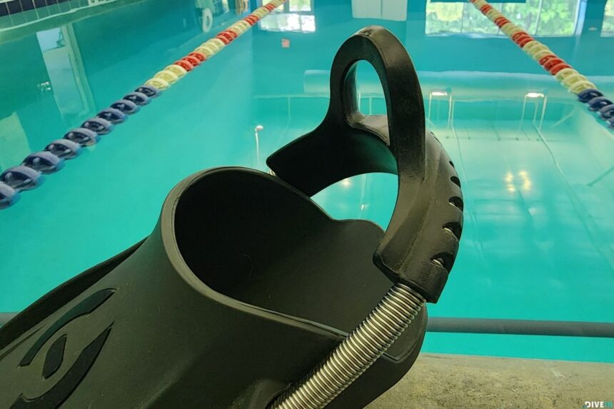 Heel strap and pull tab of a Hollis F1 LT fin propped on a poolside edge with lane ropes behind