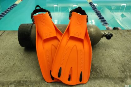 Orange ScubaPro GO Sport Gorilla fins propped against a scuba tank on a pool deck