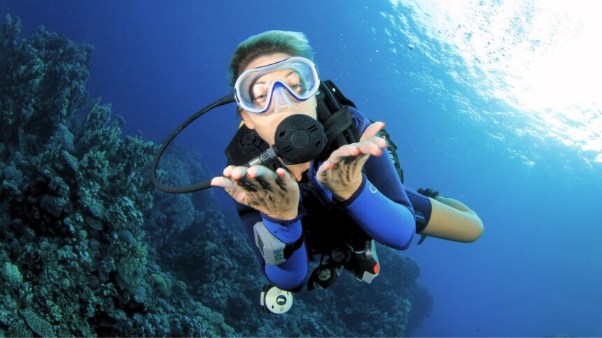 Scuba diver in a blue wetsuit hovering above a reef and gesturing toward the camera