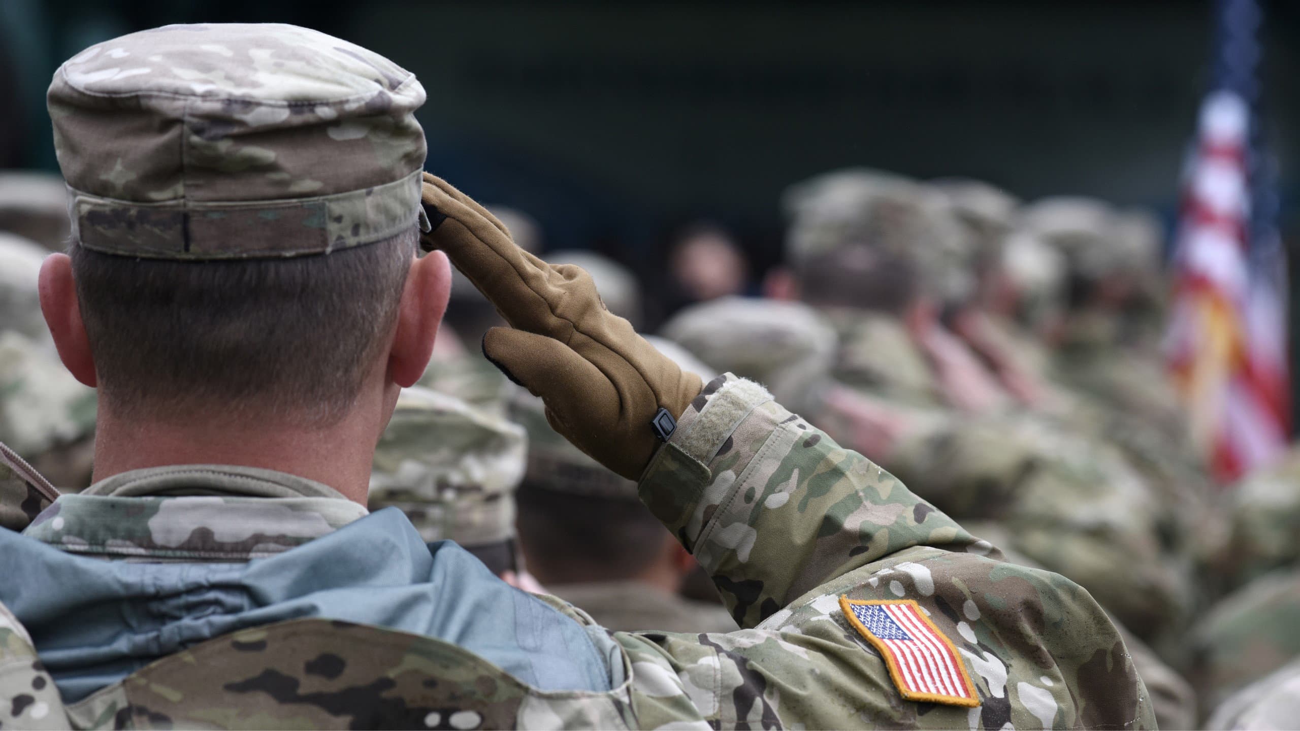 US soldiers in camouflage saluting in formation with an American flag behind them