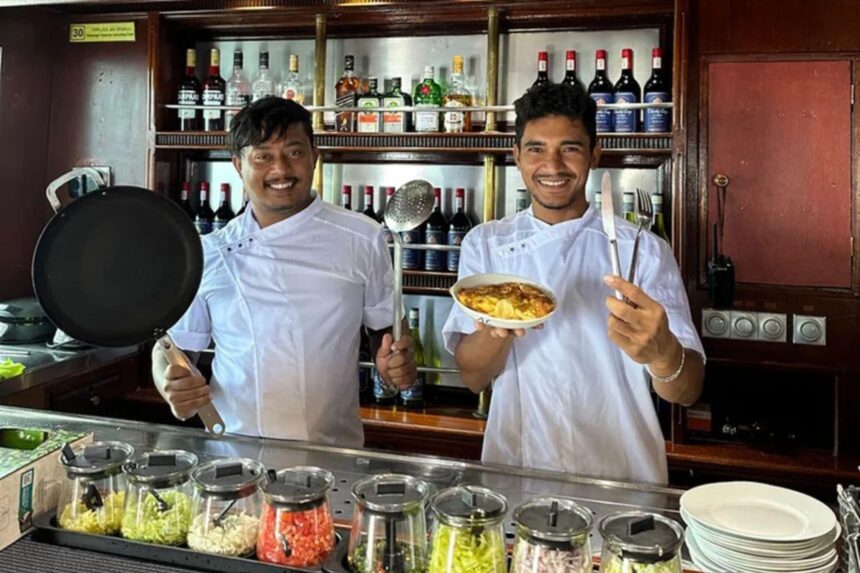 Two smiling chefs presenting a plated dish behind the galley counter on the Amba