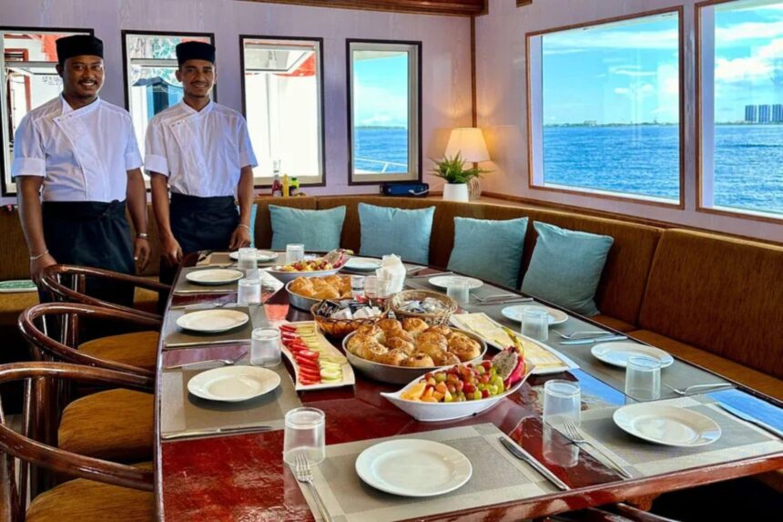 Two crew members in chef uniforms standing beside a table set with breakfast on the Amba