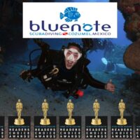 Scuba diver smiling through a reef swim-through beneath the Blue Note Scuba Cozumel logo and awards row