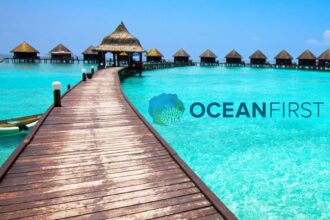 Wooden dock leading to overwater bungalows on a turquoise lagoon, overlaid with the Ocean First logo