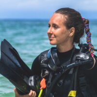 Scuba diver on a beach holding fins, her braided hair secured with a colorful Rip Tie