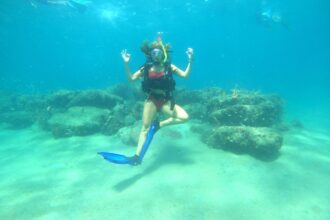 Diver striking a yoga tree pose underwater, giving the OK signal with both hands over a reef