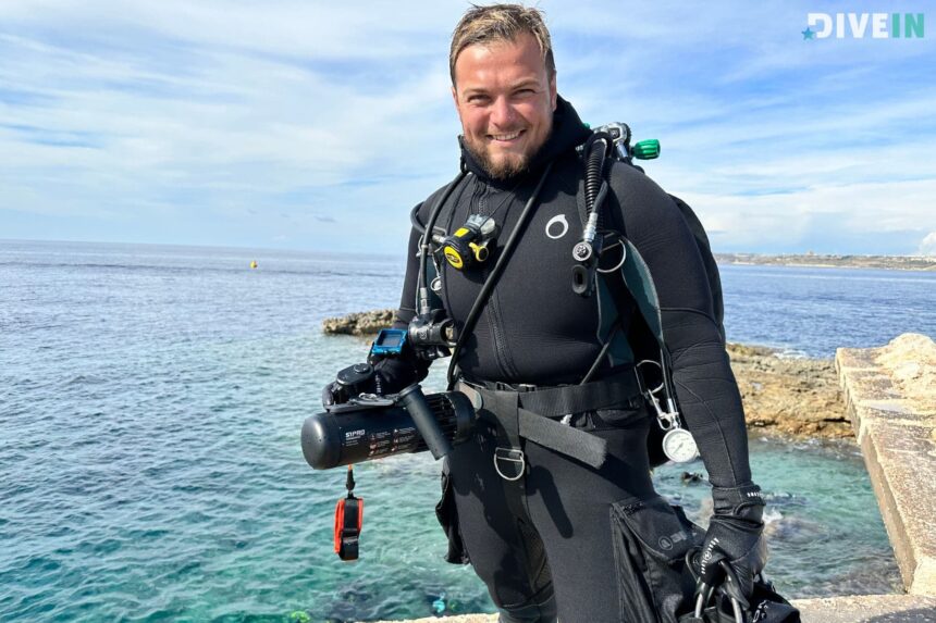 DIVEIN tester in a wetsuit on a rocky shore carrying scuba gear and a LEFEET S1 Pro before a dive