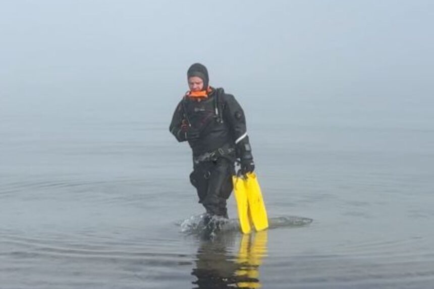 DIVEIN tester walking out of flat, foggy water in the Hollis DX-300X drysuit with yellow fins in hand