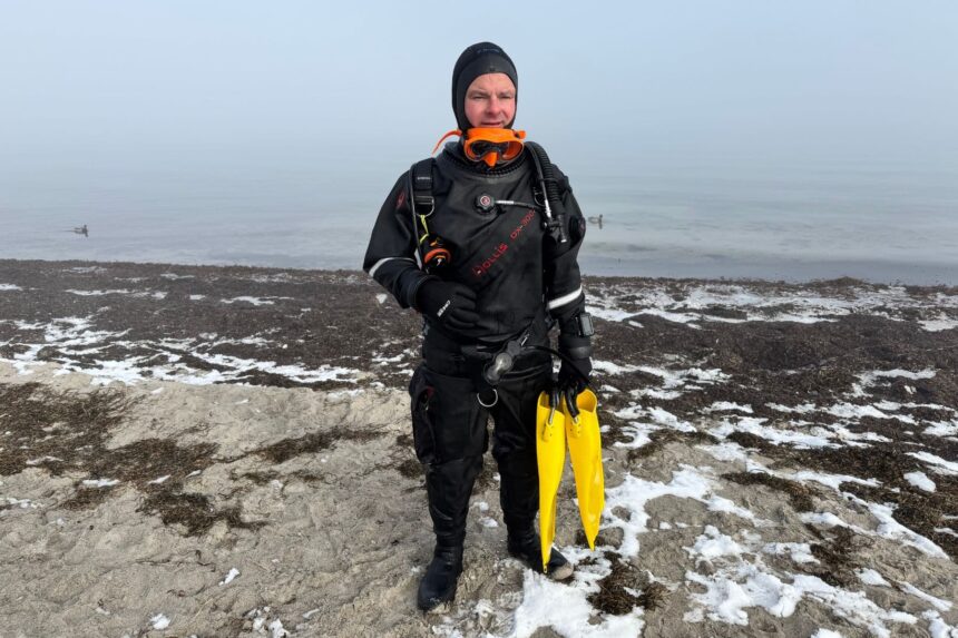 Hollis DX-300X Drysuit On Shore DIVEIN tester fully kitted in the Hollis DX-300X drysuit on a snowy shore, yellow fins in hand