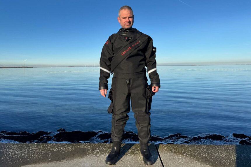 Hollis DX-300X Front View On Test Day DIVEIN reviewer wearing the Hollis DX-300X drysuit on a sunlit shore with wind turbines behind