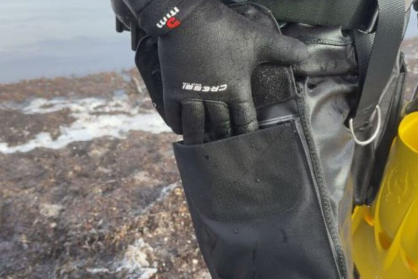 Drysuit Thigh Pocket In Use Gloved hand opening a telescopic drysuit thigh pocket, with yellow fins carried alongside