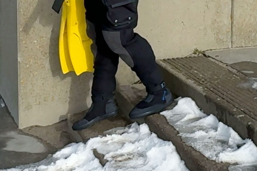 Bare Sentry Tech Drysuit Boots Attached drysuit boots on the Bare Sentry Tech, stepping across snow covered concrete steps with fins under one arm
