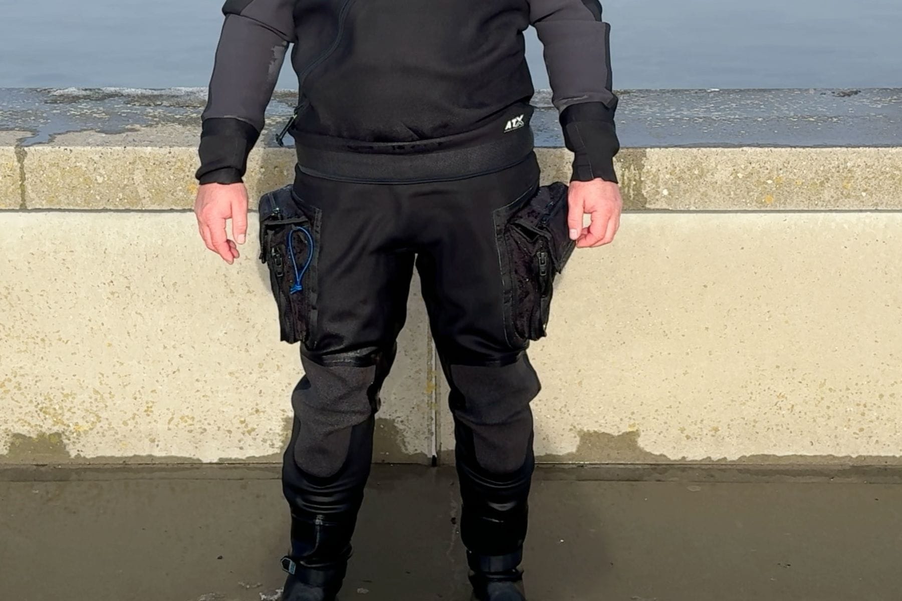 Tech Drysuit Thigh Pockets And Knees Lower half of a tech drysuit showing both thigh pockets and reinforced knees against a seawall