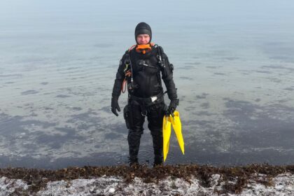 DIVEIN tester standing in shallow water in the BARE X-Mission Evolution drysuit, yellow fins in hand