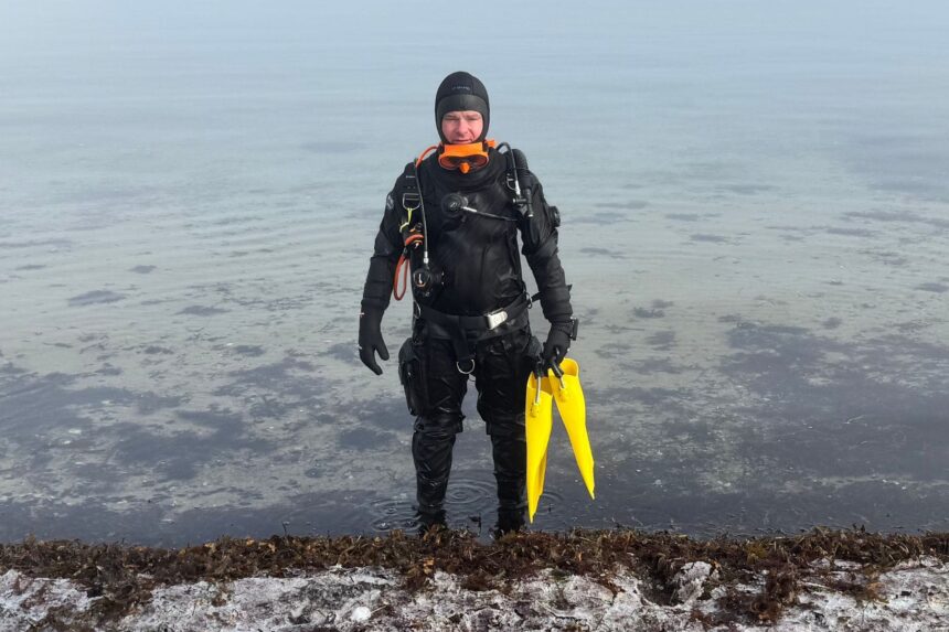 BARE X-Mission Evolution Drysuit Front View DIVEIN tester standing in shallow water in the BARE X-Mission Evolution drysuit, yellow fins in hand