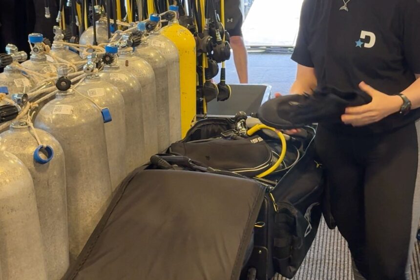 DIVEIN tester lifting fins from the packed Cressi Moby 7 dive bag beside filled tanks on a dive boat