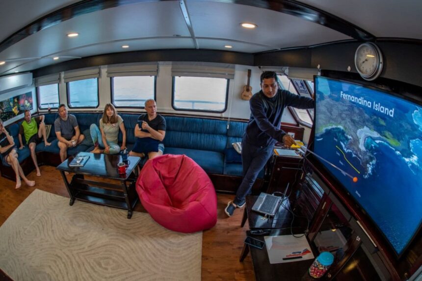 Guests in the lounge listen to a briefing with an island route shown on the TV screen