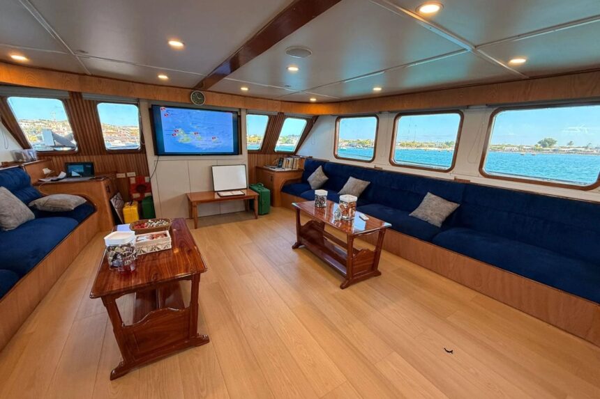 Onboard saloon with blue sofas and a wall-mounted screen showing a dive site map