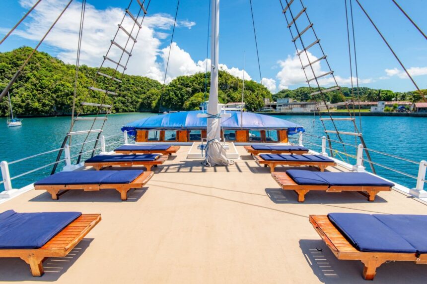 Cushioned sun loungers on deck between the schooner's rigged masts and rope ladders