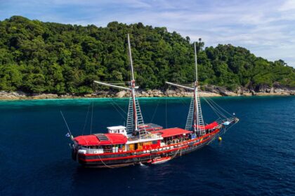 The Thailand Master liveaboard, a red-sailed traditional-style boat, anchored near a jungle coast