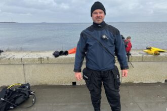 DIVEIN tester wearing the Fourth Element Argonaut 3.0 drysuit at a grey, cold-water shore dive site