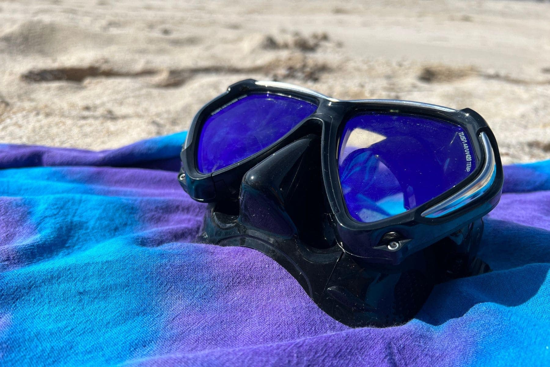 TUSA Paragon twin-lens mask on a blue beach towel, lenses tinted violet in the sun