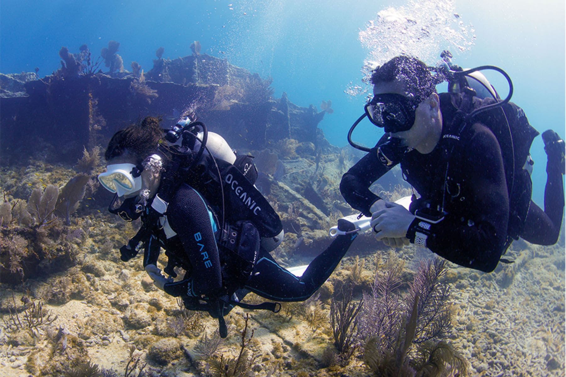 two divers explore a shallow shipwreck reef, one wearing an Oceanic-branded wetsuit and BCD