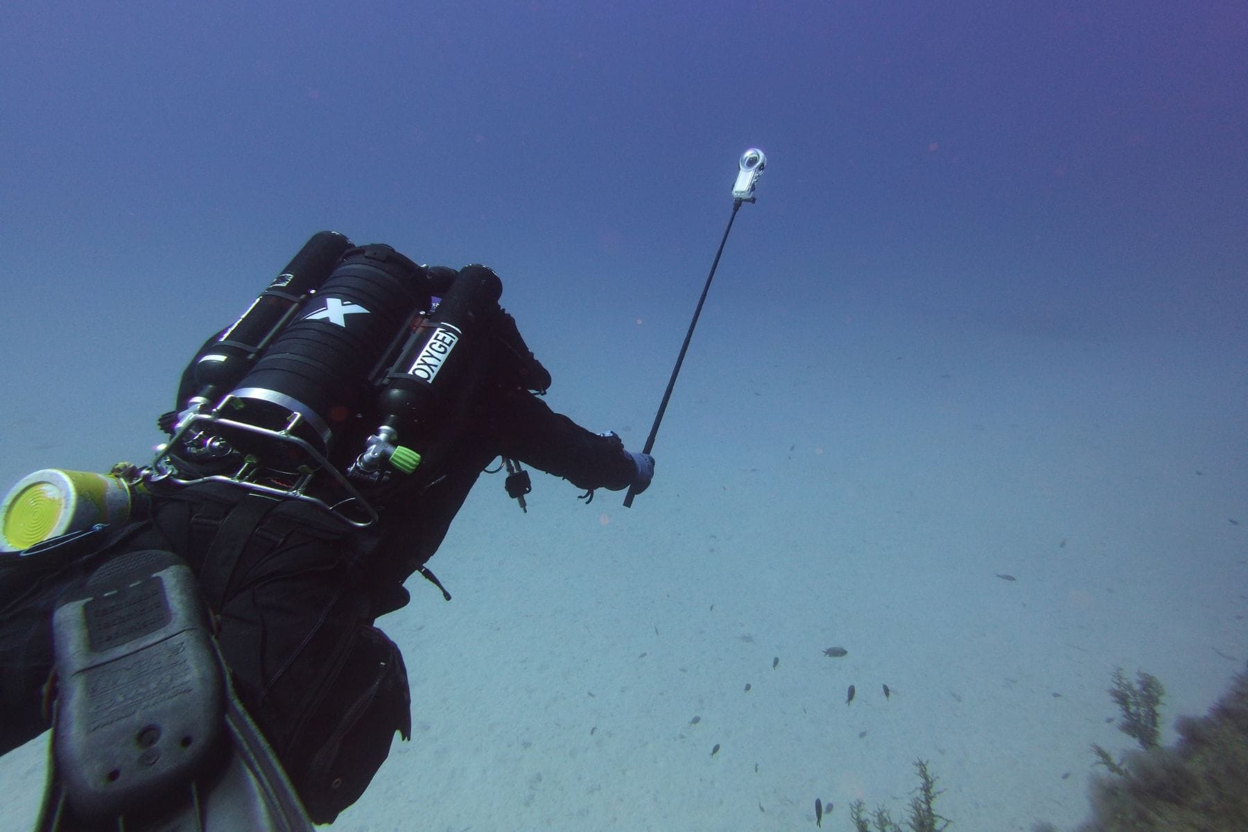 rebreather diver holding an action camera on an extension pole above a sandy seabed