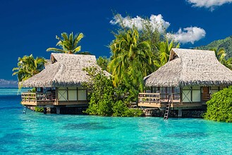 Two thatched overwater bungalows on stilts above a bright turquoise lagoon fringed with palms