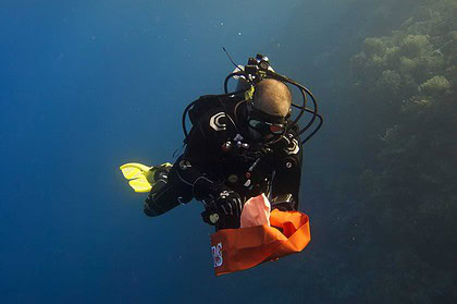 Diver reaching up to inflate an orange DSMB at the surface with dive boats nearby