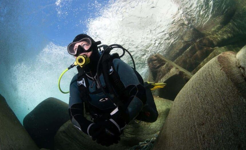 Scuba diver in a black wetsuit kneeling among boulders in clear, sunlit shallow water