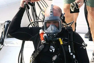 Commercial diver in a full face mask and tangled air hoses being helped into gear on a boat deck