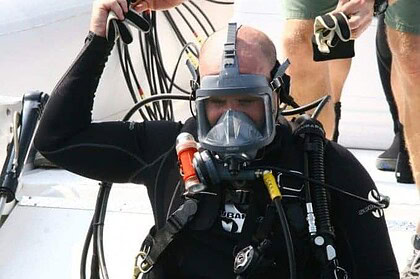 Commercial diver in a full face mask and tangled air hoses being helped into gear on a boat deck