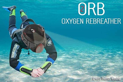 A diver wearing the ORB oxygen rebreather helmet floating horizontally over a sandy seabed